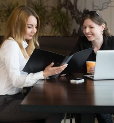 Two professional women discussing documents at a table in a modern office setting. One woman is holding a black folder while the other is smiling, with a laptop and coffee cup in front of her.