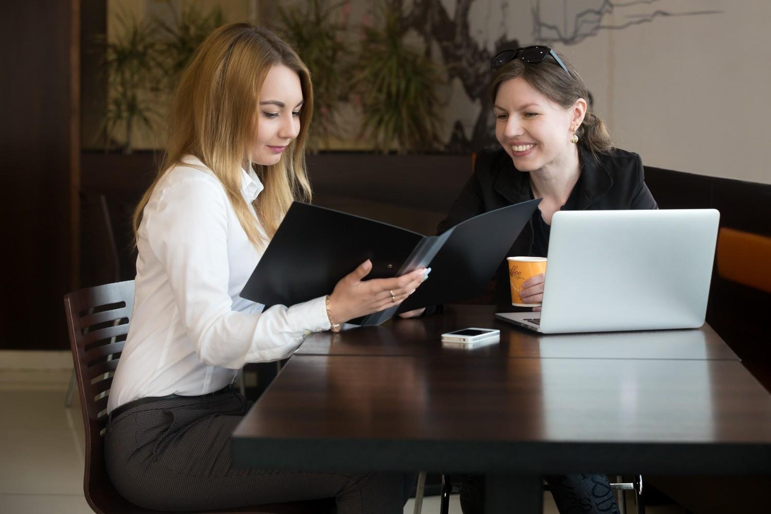 Two professional women discussing documents at a table in a modern office setting. One woman is holding a black folder while the other is smiling, with a laptop and coffee cup in front of her.