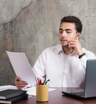 A professional man reviewing H-1B visa stamping documents while talking on the phone, with a laptop and books on his desk.