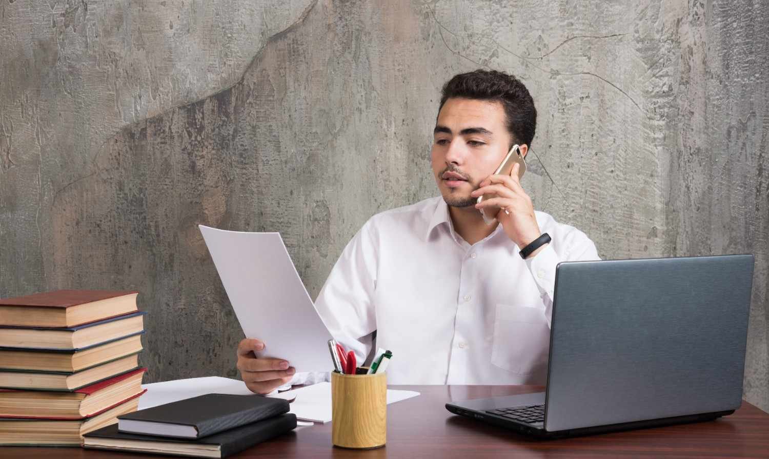 A professional man reviewing H-1B visa stamping documents while talking on the phone, with a laptop and books on his desk.