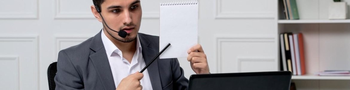 A professional man in a suit wearing a headset, sitting at a desk with a laptop, holding up a blank notepad and pointing at it with a pen, likely explaining something during a virtual meeting.
