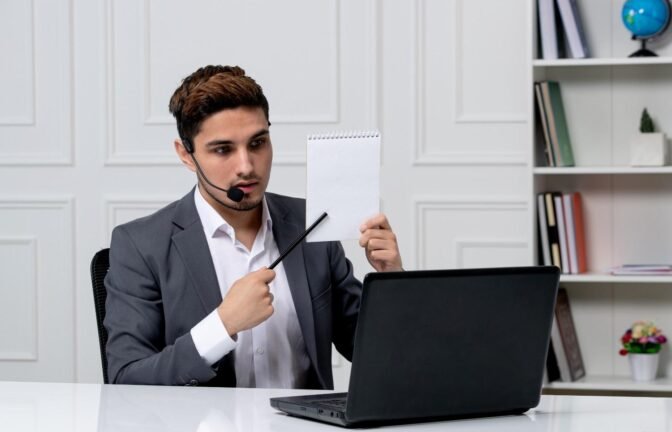 A professional man in a suit wearing a headset, sitting at a desk with a laptop, holding up a blank notepad and pointing at it with a pen, likely explaining something during a virtual meeting.