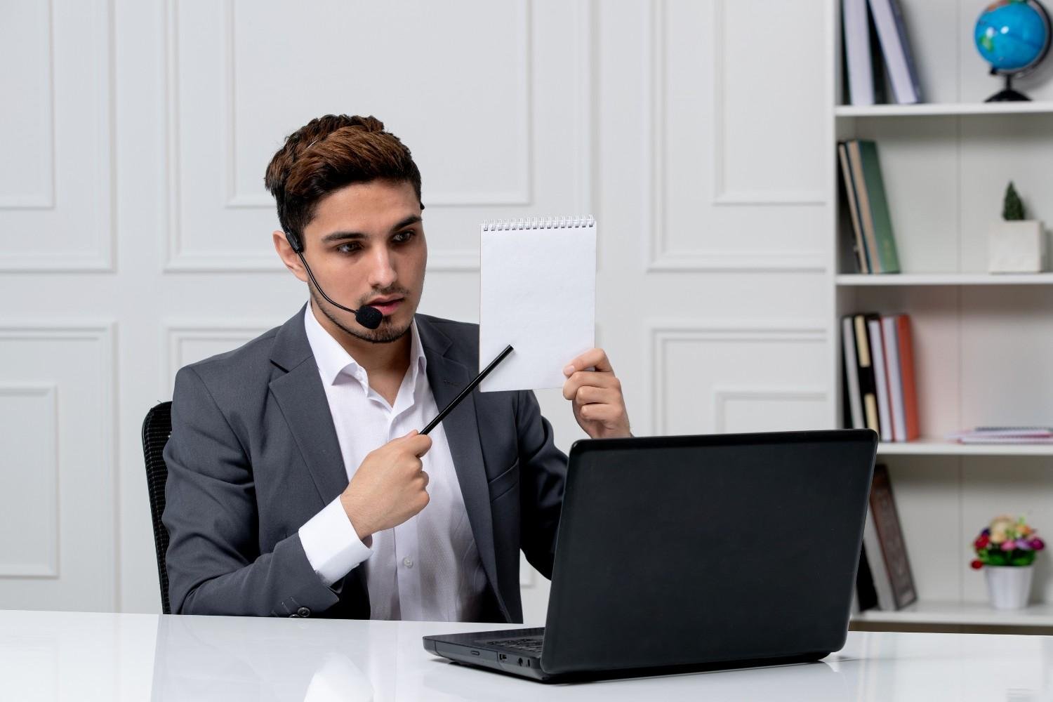 A professional man in a suit wearing a headset, sitting at a desk with a laptop, holding up a blank notepad and pointing at it with a pen, likely explaining something during a virtual meeting.
