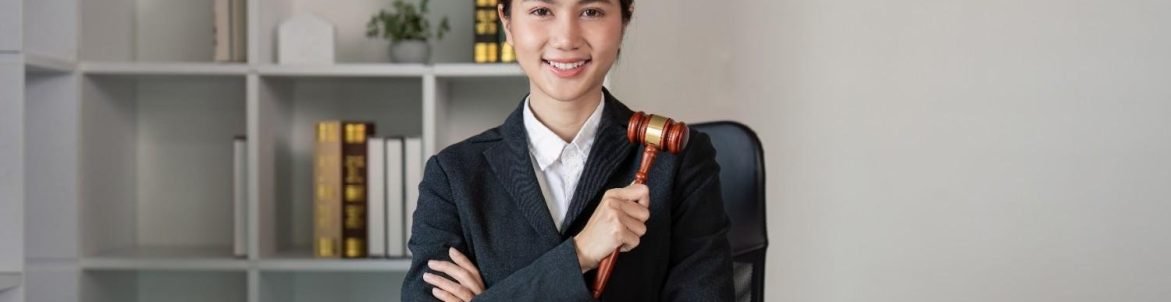 Smiling Virtual Legal Assistant in 2025 holding a gavel, sitting at a modern law office desk with a laptop, legal scales, and documents.