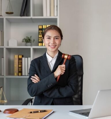 Smiling Virtual Legal Assistant in 2025 holding a gavel, sitting at a modern law office desk with a laptop, legal scales, and documents.